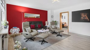 A modern living room with a red accent wall, a sofa, two armchairs, a coffee table, and a small side table with books. - FeWo Galliet 1: 87sqm, 2 BR, balcony, quiet & close to trade fair (NRW)