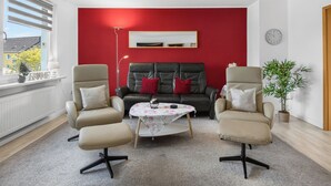 A living room with two armchairs, a sofa, a small round table, a red accent wall, a clock, and a window with blinds. - FeWo Galliet 1: 87sqm, 2 BR, balcony, quiet & close to trade fair (NRW)