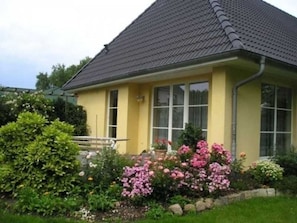 Ein gelbes Gebäude mit grauem Dach, umgeben von einem Garten mit rosa Blumen.