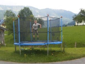 Een trampoline in een open omgeving, met een grasveld en bergen op de achtergrond.