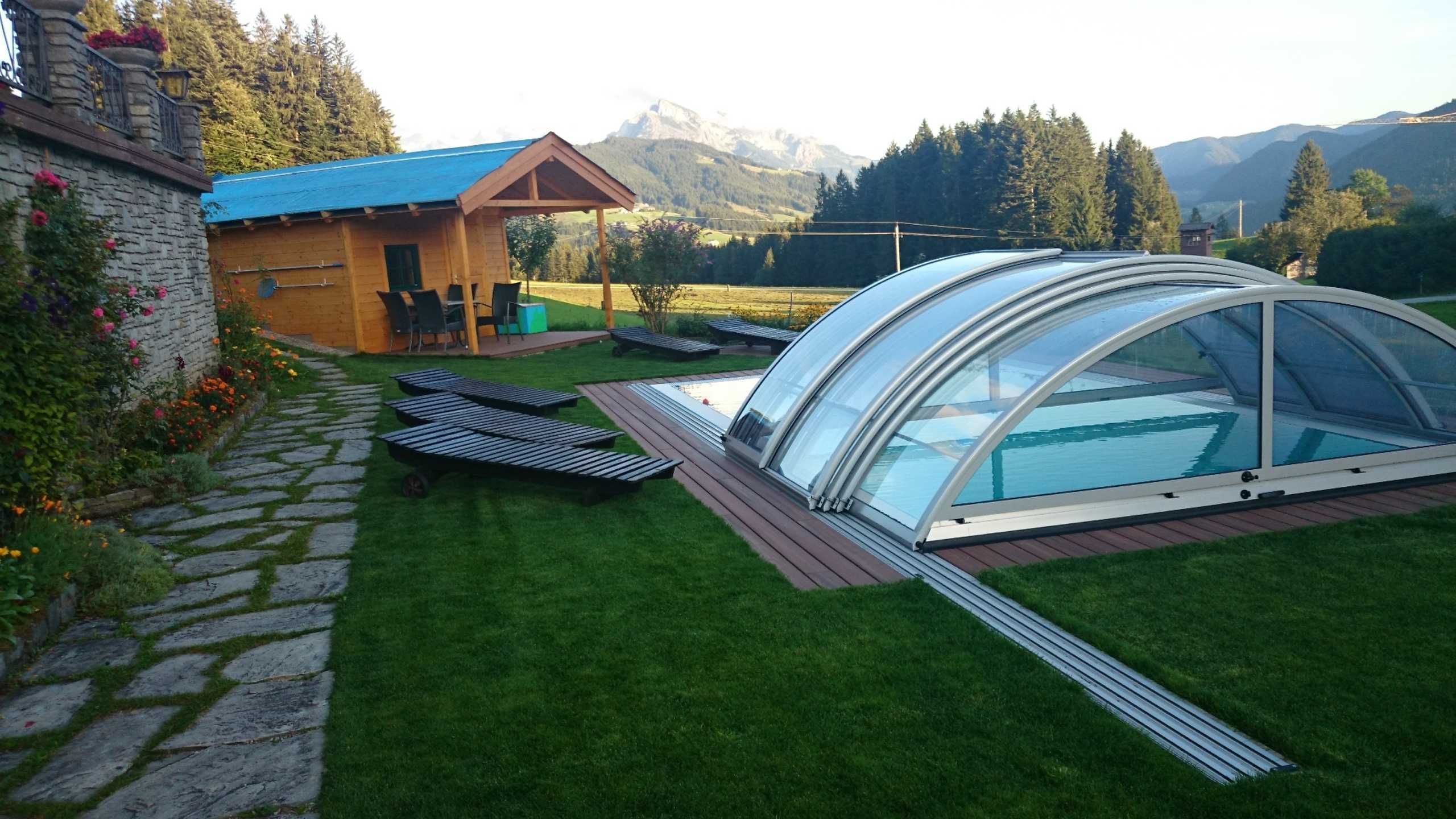 Een zwembad met glazen omheining, een houten terras en een klein gebouw met blauw dak.