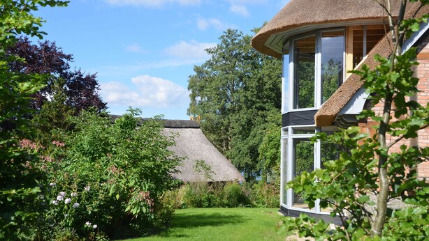 A thatched-roof house with a large window and a garden with various plants and flowers. - Duck's Nest - Thatched Aspen House (Grebin)