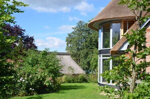 A thatched-roof house with a large window and a garden with various plants and flowers. - Duck's Nest - Thatched Aspen House (Grebin)