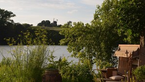 A riverside setting with a wooden deck, a rustic windmill in the background, and lush greenery. - Duck's Nest - Thatched Aspen House (Grebin)