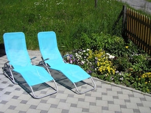 Zwei blaue Lounge-Stühle auf einer gepflasterten Terrasse mit einem Garten, der verschiedene Blumen und einen Holzzaun im Hintergrund zeigt.