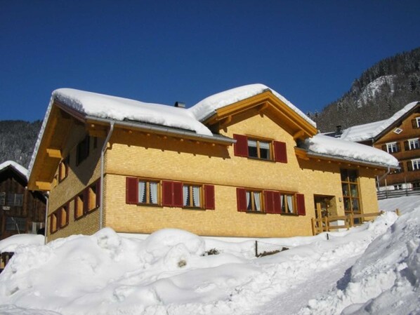 A yellow building with red shutters, surrounded by snow and mountains.