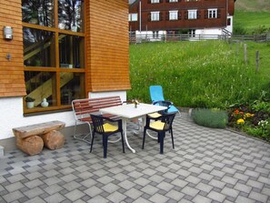 Une terrasse avec une table et des chaises, un banc en bois, et une vue sur un champ verdoyant et un bâtiment en arrière-plan.
