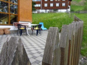 Ein Holzzaun, ein Schachbrettmuster auf dem Boden, Gartenmöbel und ein Gebäude im Hintergrund.