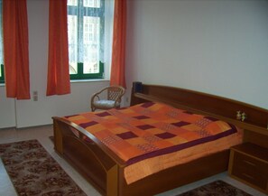 Quarto com cama de madeira, uma cadeira, mesa de cabeceira e janela com cortinas.