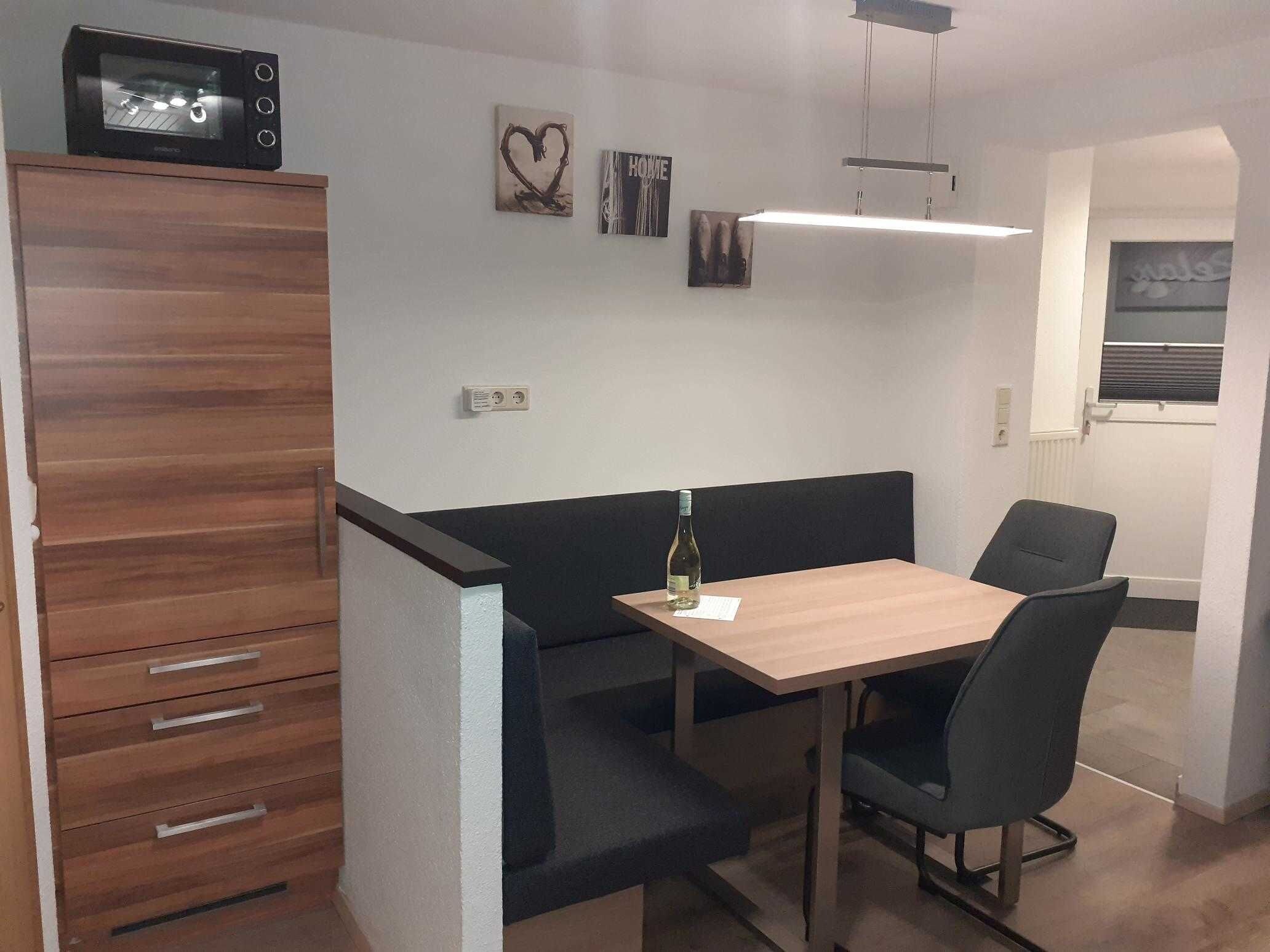 A compact room with a wooden cabinet, a table with two chairs, and a bench. There are wall-mounted electrical outlets and a small window with blinds.