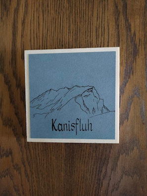 Tarjeta con ilustración de una montaña y el texto 'Kanisfluh' sobre un fondo de madera.