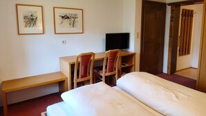 Ein Hotelzimmer mit einem Bett, einem Schreibtisch mit Computer, zwei Stühlen, einer Bank und zwei gerahmten Bildern an der Wand.