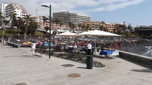 Eine Strandpromenade mit Außenbestuhlung und Sonnenschirmen, mit Blick auf einen belebten Strand und das Meer.
