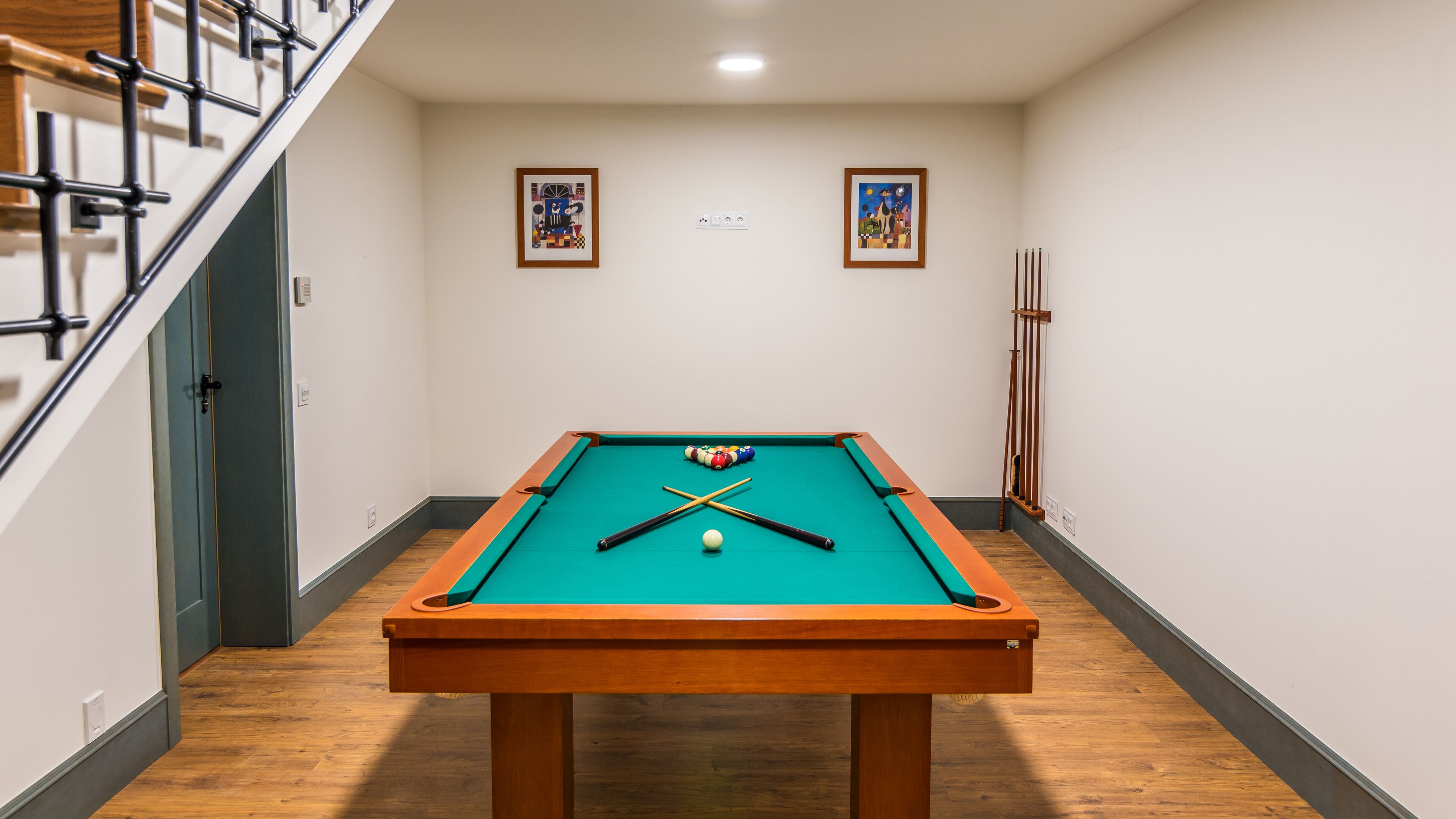 Une table de billard se trouve dans un couloir, le sol est recouvert de parquet, un escalier est situé à gauche, et deux œuvres d’art encadrées ornent les murs.