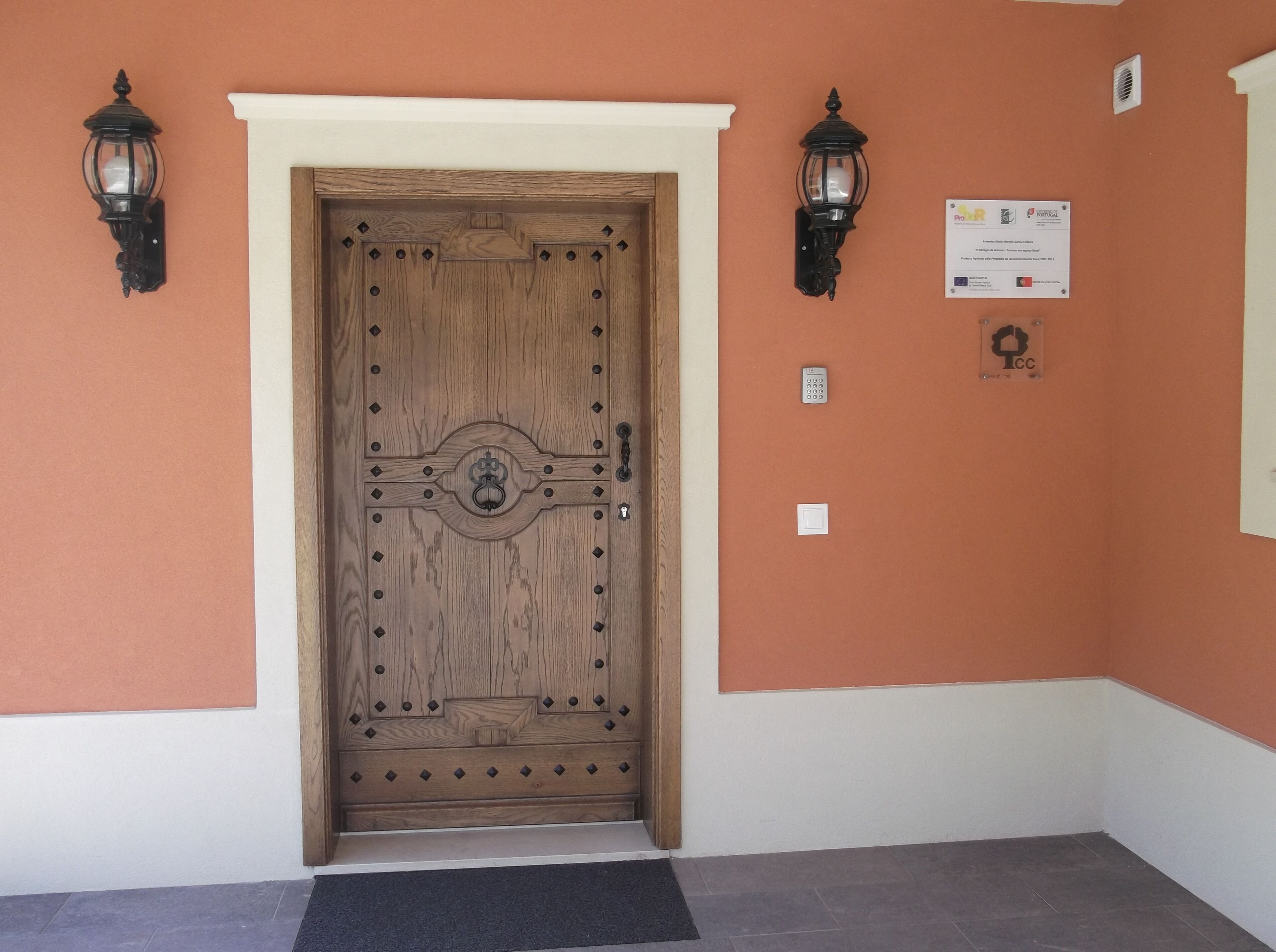 Una puerta de madera con un tirador y cerradura decorativos, flanqueada por dos faroles montados en la pared. Una placa con logotipos y texto es visible en la pared a la derecha de la puerta.