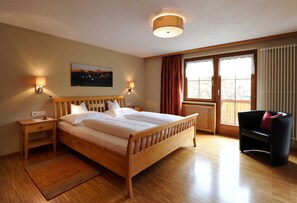 A bedroom with a wooden bed, nightstands, a wall-mounted lamp, a window with curtains, and a view of the outdoors.