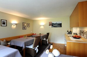A dining area with tables and chairs, a kitchen counter with a fruit bowl, and wall-mounted lights.