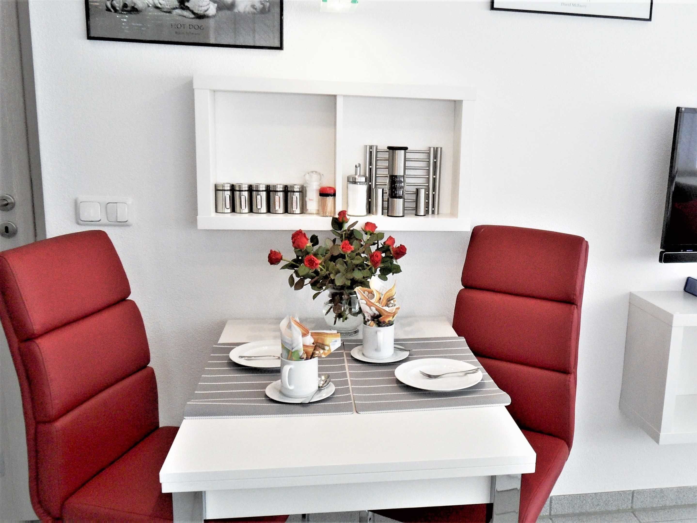 A dining area with a table set for two, red chairs, a vase with red roses, and a white shelf above the table with kitchen items.