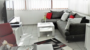 Ein modernes Wohnzimmer mit einem grauen Sofa, einem weißen Couchtisch und einem roten Kissen. Ein Fernseher steht auf einem Lowboard, ein kleiner Tisch mit einer Vase und ein Fenster mit Jalousien sind ebenfalls zu sehen.