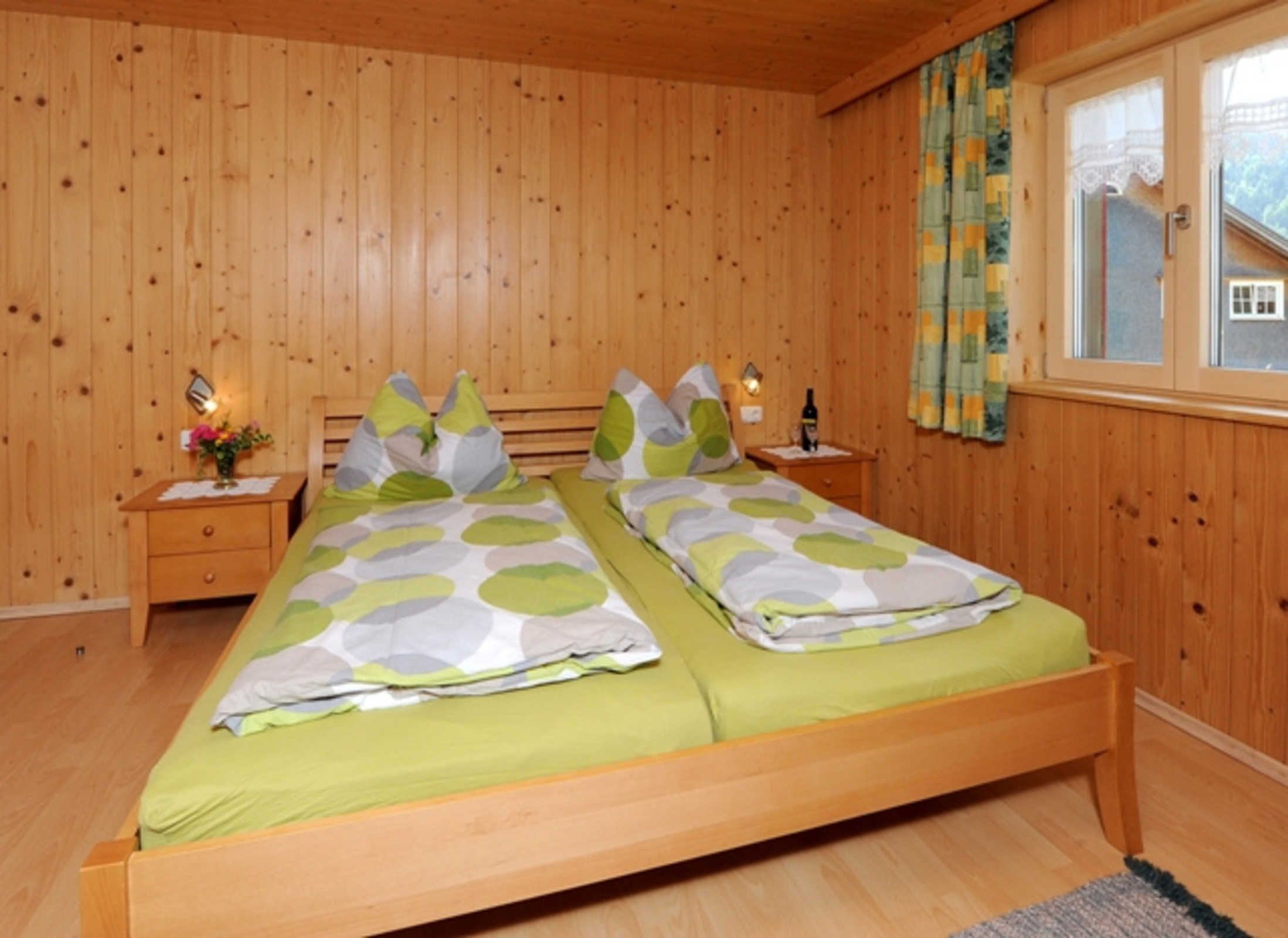 A wooden-paneled bedroom with two beds, a nightstand with a lamp, and a window with curtains.