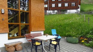 A patio with a table and chairs, a wooden bench, and a view of a green field and a building in the background. - Apartment 1 1 - Bishop Reingard (Schoppernau)