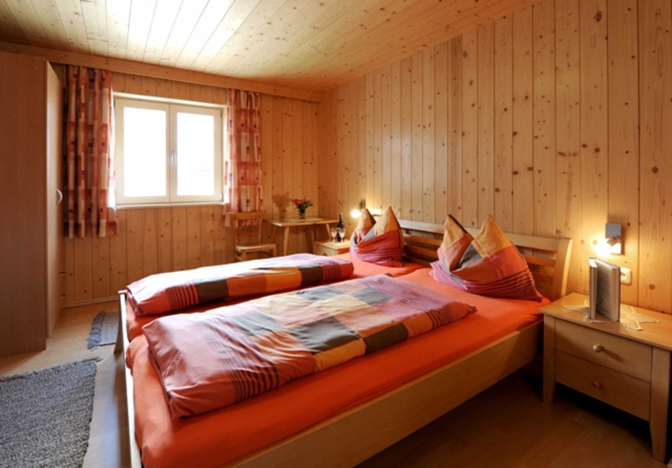 Quarto com painéis de madeira, uma cama, mesa de cabeceira, mesa com cadeiras e janela com cortinas.