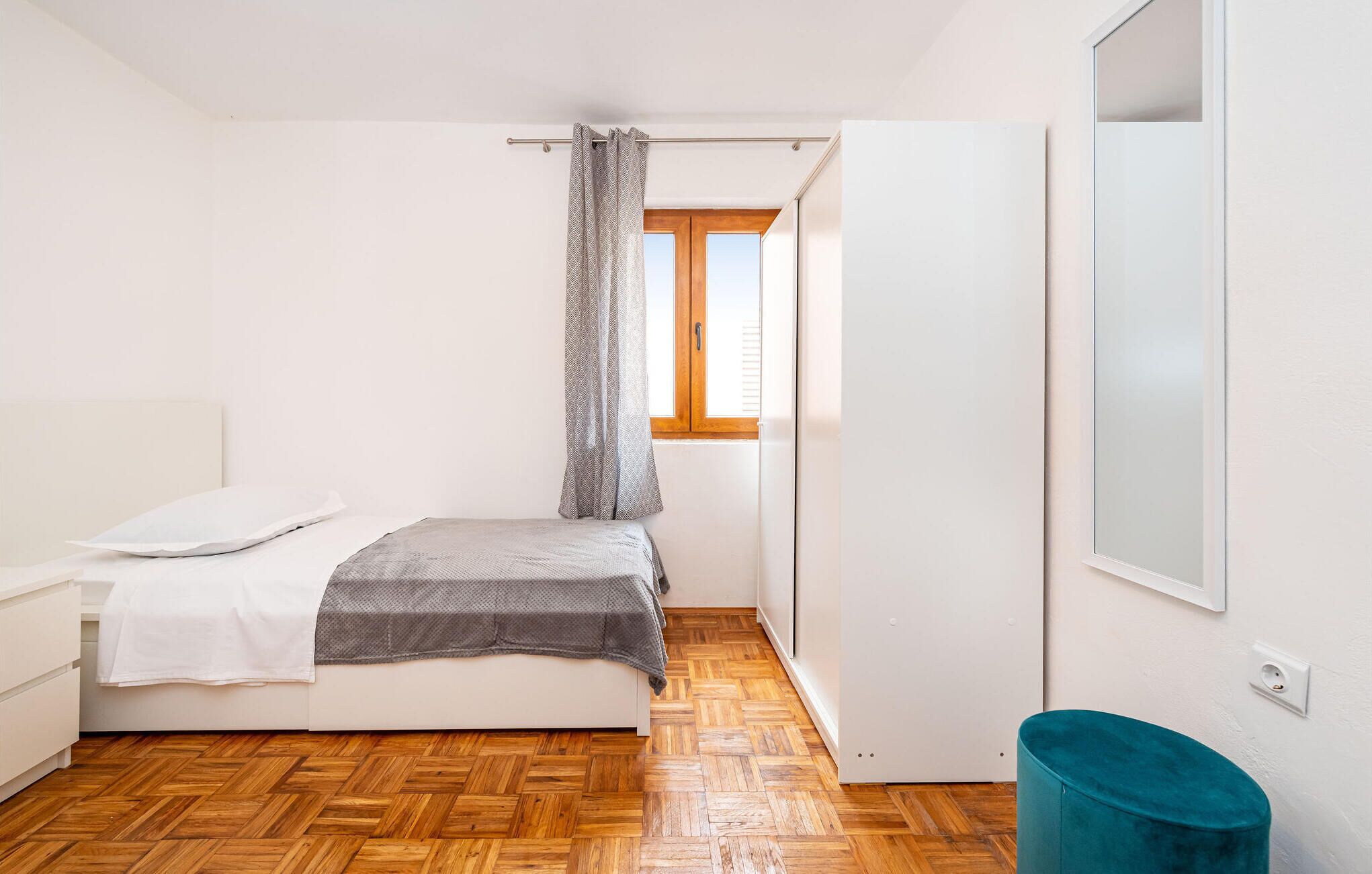 Ein Schlafzimmer mit einem Bett, einem Fenster mit Vorhängen, einem Holzboden und einem Türkischgrünfarbenen Sessel.