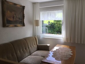 Un salon avec un canapé, une table sur laquelle est posée une nappe en dentelle, une lampe, une fenêtre avec des rideaux et un tableau accroché au mur.