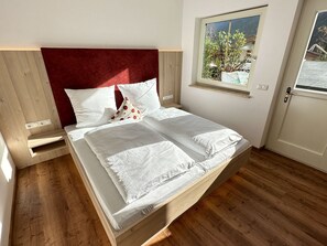Un lit simple avec un cadre en bois et une tête de lit rouge, une literie blanche et une fenêtre donnant sur un paysage verdoyant.