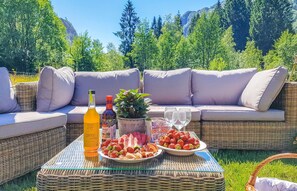 Un sofá de mimbre con cojines, una mesa de vidrio con una botella, una planta en maceta y un plato de fresas.