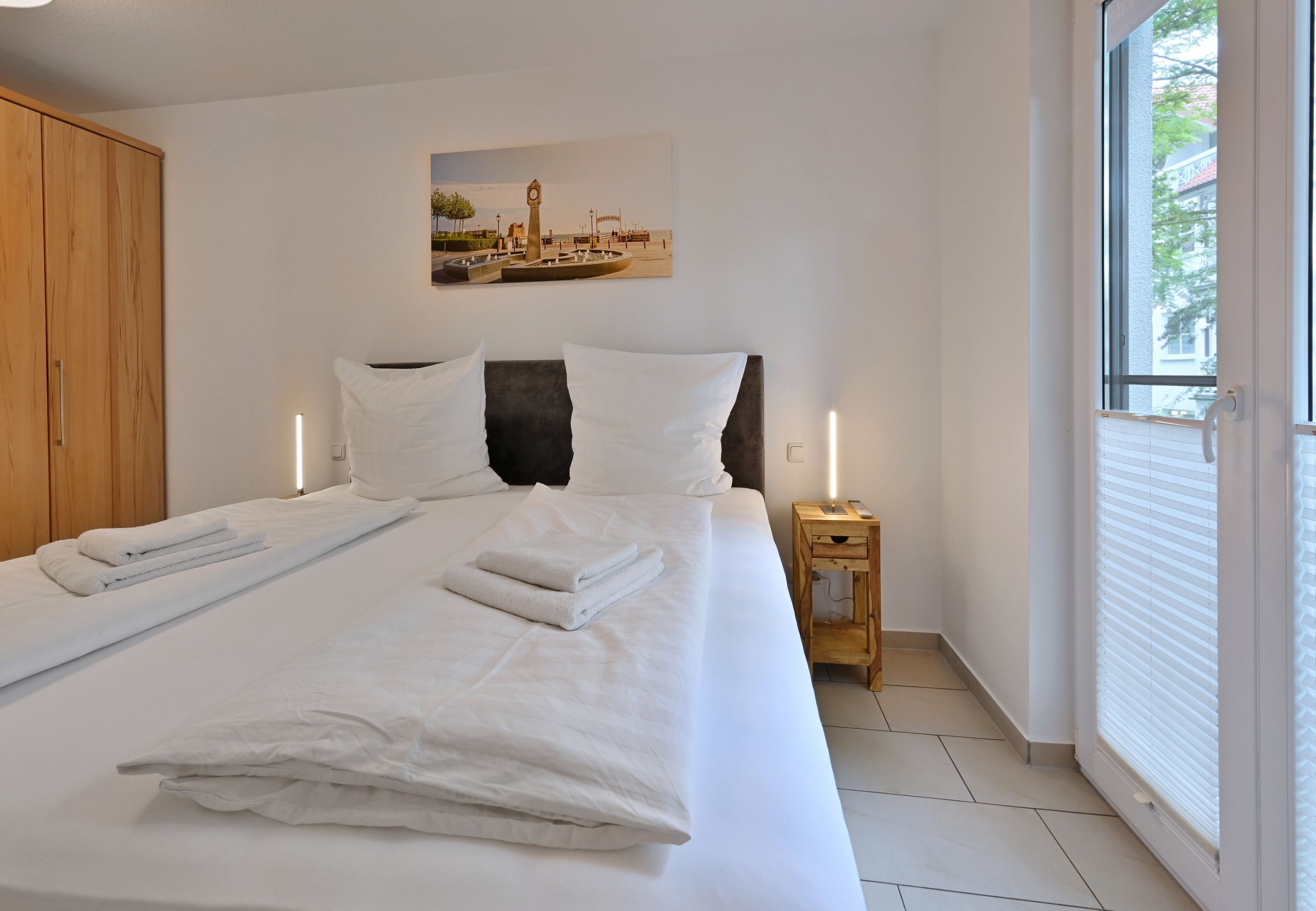 Una cama bien hecha con sábanas y toallas blancas, un armario de madera, una mesita de noche con una lámpara y un cuadro de un paisaje urbano en la pared.
