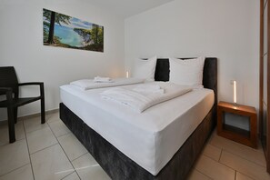 A neatly made bed with white linens and pillows, a dark headboard, a small bedside table with a lamp, and a chair.