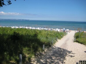 Ein Strandweg führt zu Strandstühlen und Sonnenschirmen, mit freiem Blick auf das Meer und das entfernte Land.