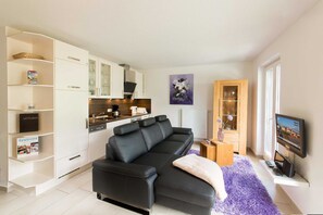 A modern living room with a black leather sofa, a kitchen area with white cabinets, a purple rug, and a television mounted on the wall.