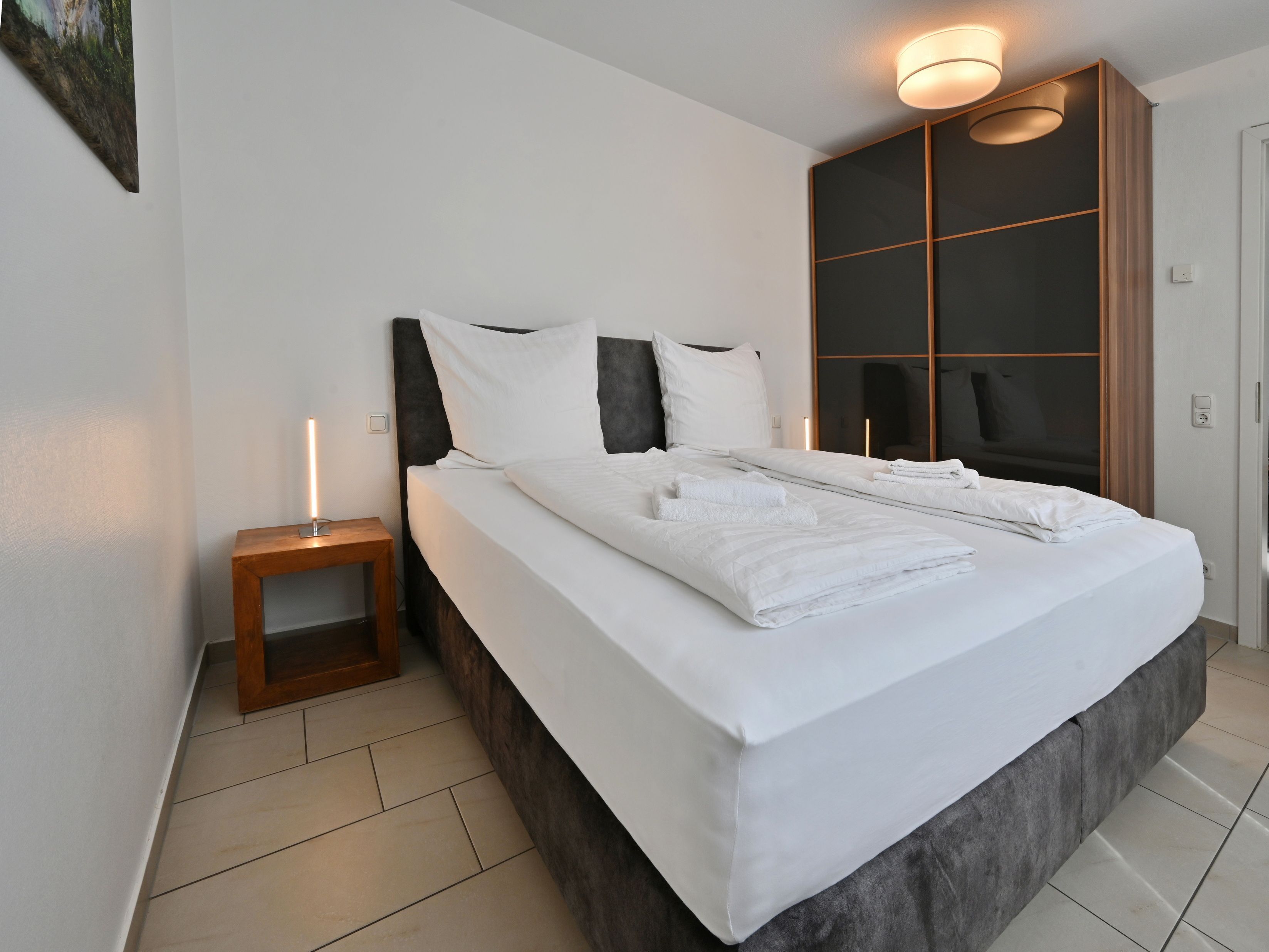Una habitación de hotel moderna con una cama grande, una mesita de noche con lámpara, un armario y un espejo.