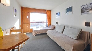 A room with a wooden table, a sofa, and a bed. There are pictures on the wall and a window with curtains.