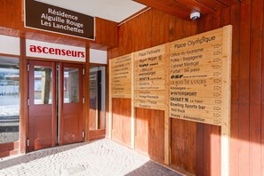 Un bâtiment en bois, sur lequel on peut lire l’inscription « ascenseurs », et de nombreux panneaux indicateurs indiquant la direction de différents lieux, comme la « Place Olympique », la « Place Patinoire » et « InterSport ».