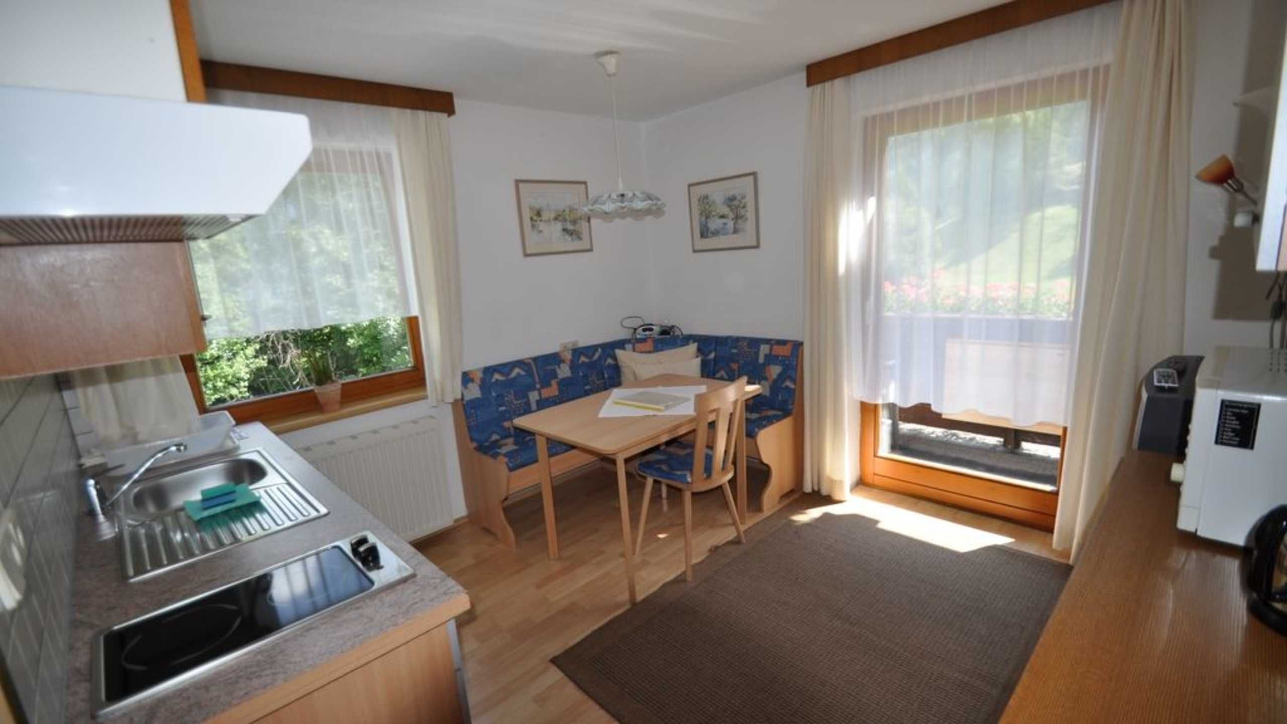 A small kitchenette with a sink and a stove, a dining area with a table and chairs, and a window with curtains.