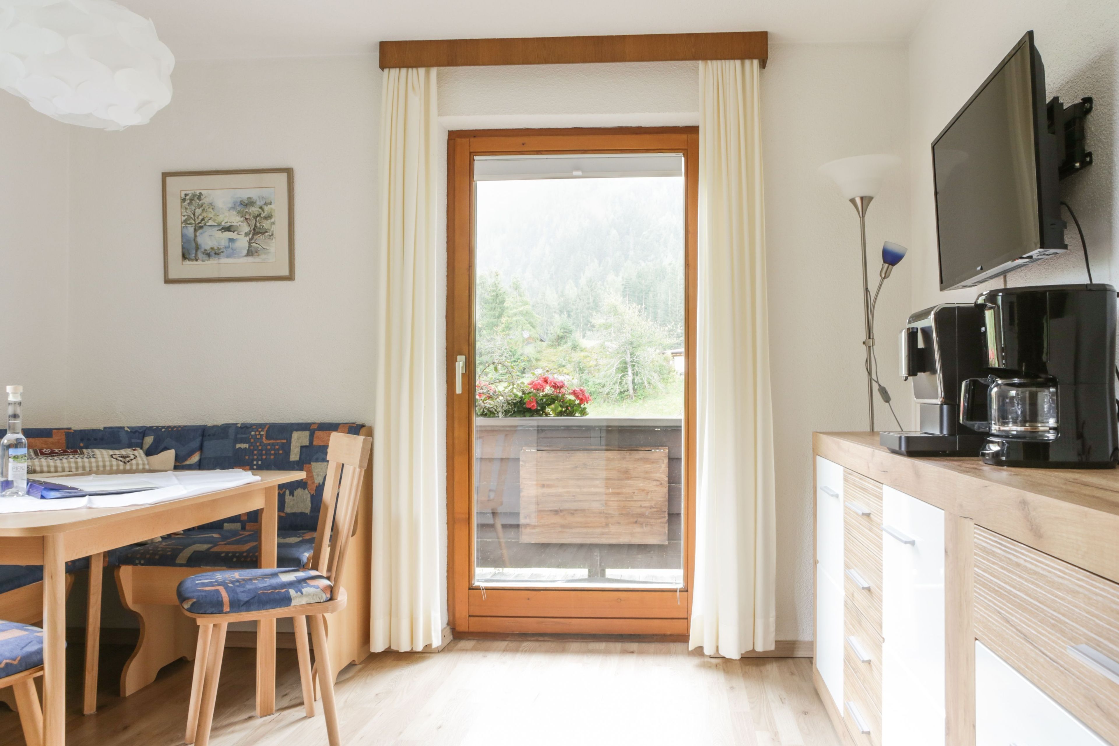Ein Zimmer mit einem Holzespeisetisch, blau gepolsterten Stühlen, einer Holzkommode, einem Fernseher und Blick auf Grün hinter einer Glastür.