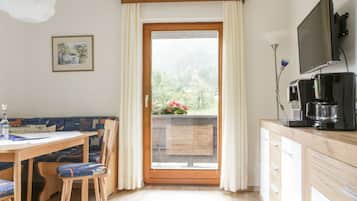 Ein Zimmer mit einem Holzespeisetisch, blau gepolsterten Stühlen, einer Holzkommode, einem Fernseher und Blick auf Grün hinter einer Glastür.