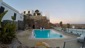 A swimming pool with a stone wall and a lifebuoy, surrounded by plants and a modern building with a stone facade. - ALTOS DE ARGUINEGUÍN (Mogán Arguineguín)