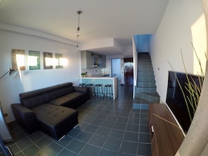 A modern living room with a sectional sofa, a dining area with stools, a kitchen, and a staircase leading to another level.