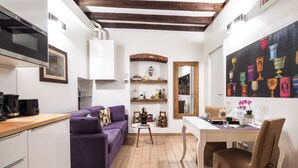 A compact living space with a kitchenette, a purple sofa, a dining table with chairs, and a wall-mounted shelf with decorative items.