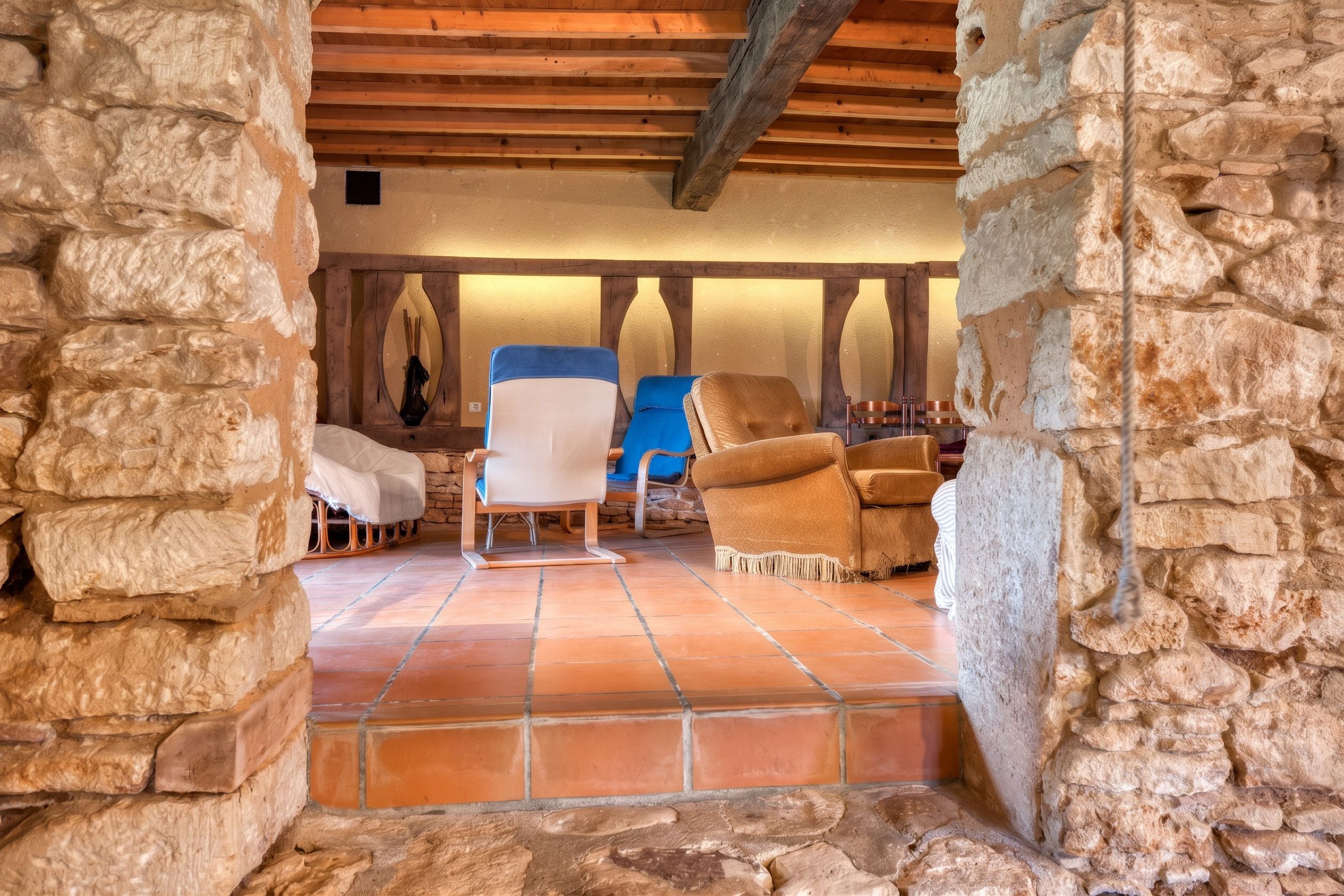 A stone-walled room with a wooden ceiling, a tiled floor, and two upholstered chairs.