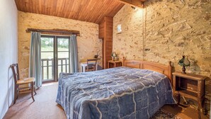 Un dormitorio rústico con pared de piedra, techo de madera, cama con colcha azul estampada, mesita de noche de madera y una silla.