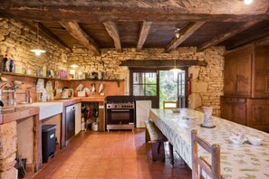 Una cocina rústica con paredes de piedra, vigas de madera, suelo de baldosas y una zona de comedor con banco y mesa.