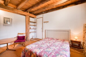 Een slaapkamer met een houten plafond, een bed met een bloemenbedekking, een stoel, een nachtkastje met een lamp, en een klein plankje met decoratieve objecten.