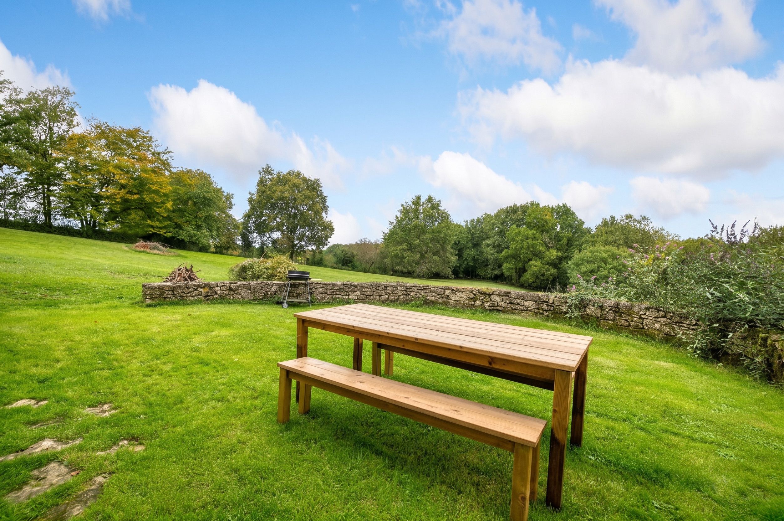 Une table de pique-nique en bois, avec des bancs, se trouve dans un champ herbeux, avec des murs de pierre et des arbres en arrière-plan.