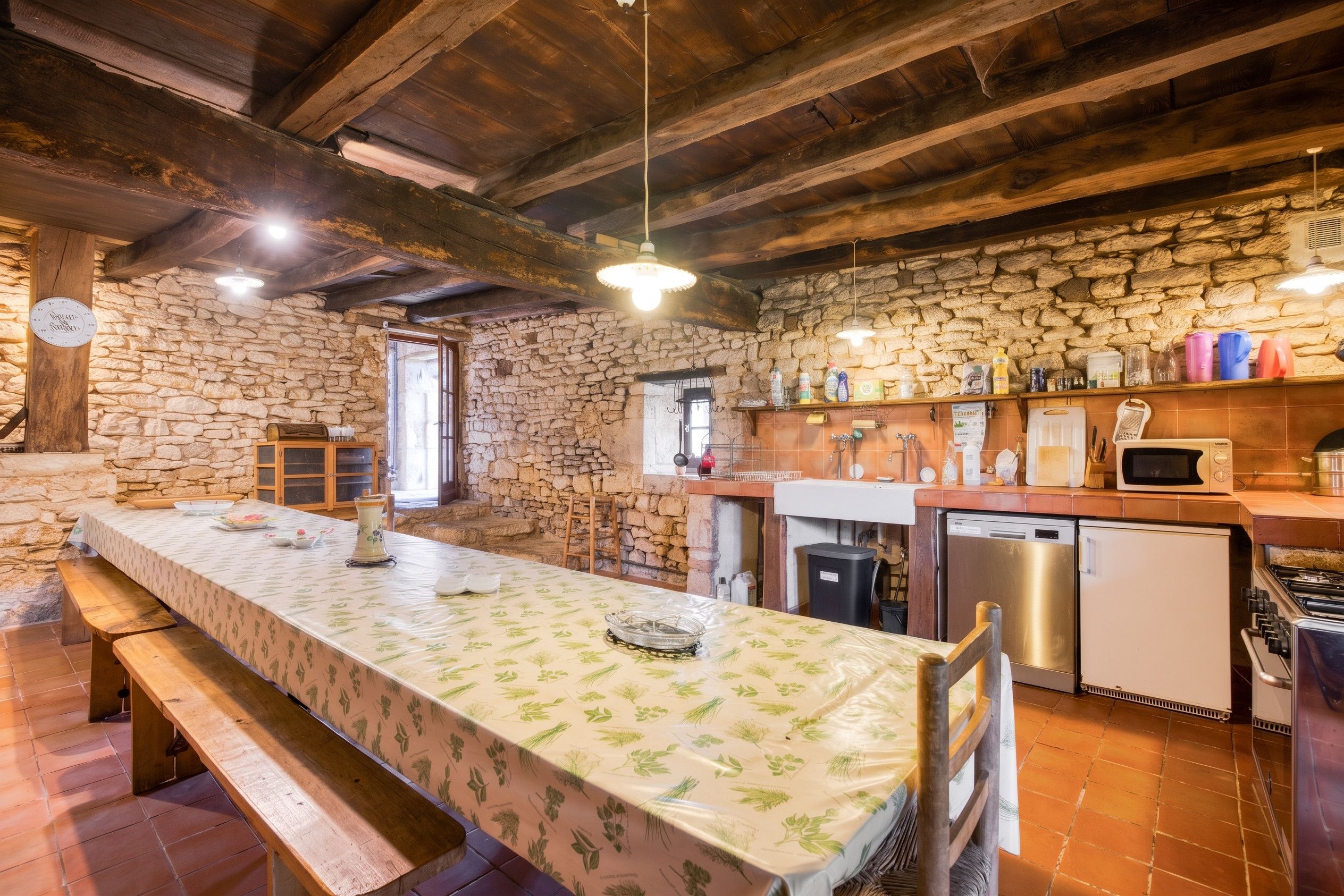 A rustic kitchen with a stone wall, wooden beams, a long dining table, and modern appliances.