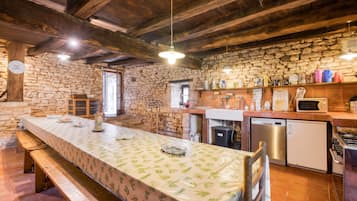A rustic kitchen with a stone wall, wooden beams, a long dining table, and modern appliances.
